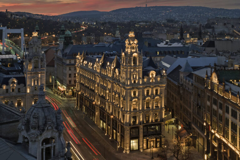 Aerial view of a historic European city center at dusk, with ornate, illuminated buildings lining a busy street and light trails from passing cars.
