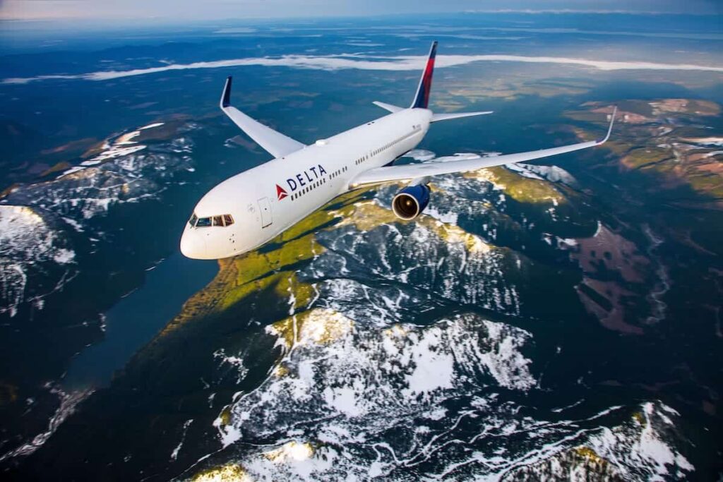 Delta airliner cruising high above snow‑capped mountains and water.