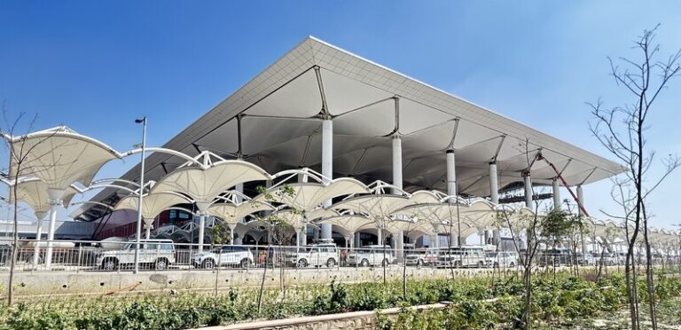 Modern transportation terminal with large curved white canopies and columns, cars parked along the front under a clear blue sky.