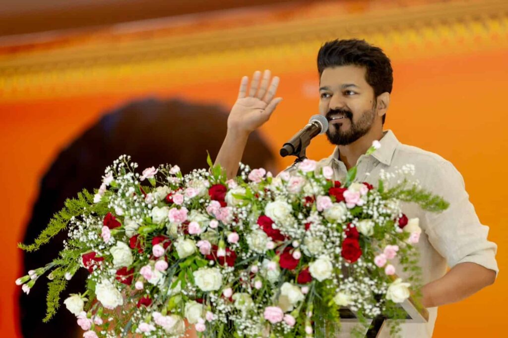 Speaker at a podium with a large flower arrangement, raising his right hand while addressing an audience.