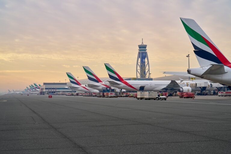 Row of Emirates airplanes parked at gates along a sunlit airport tarmac, with a distant control tower under a warm sky.