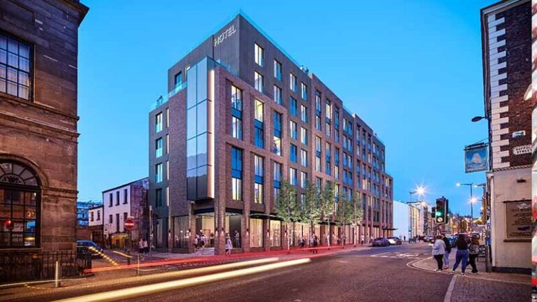 Modern brick hotel building on a city street at dusk, with pedestrians and storefront lights.