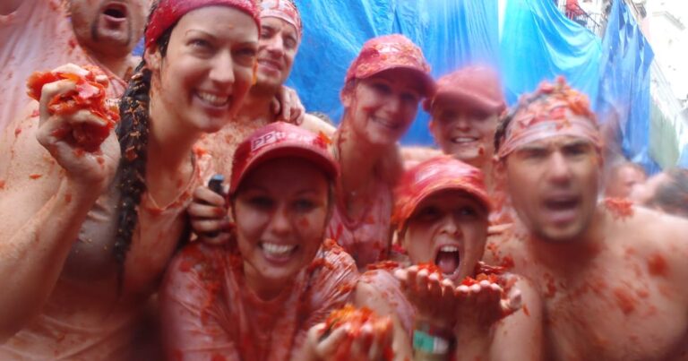 Group of smiling people covered in red tomato sauce, posing together at a tomato festival backdrop.