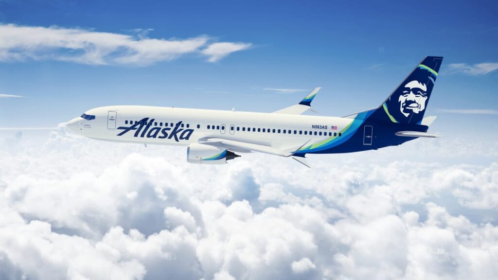 Airline passenger jet with Alaska livery flying above white clouds against a bright blue sky.