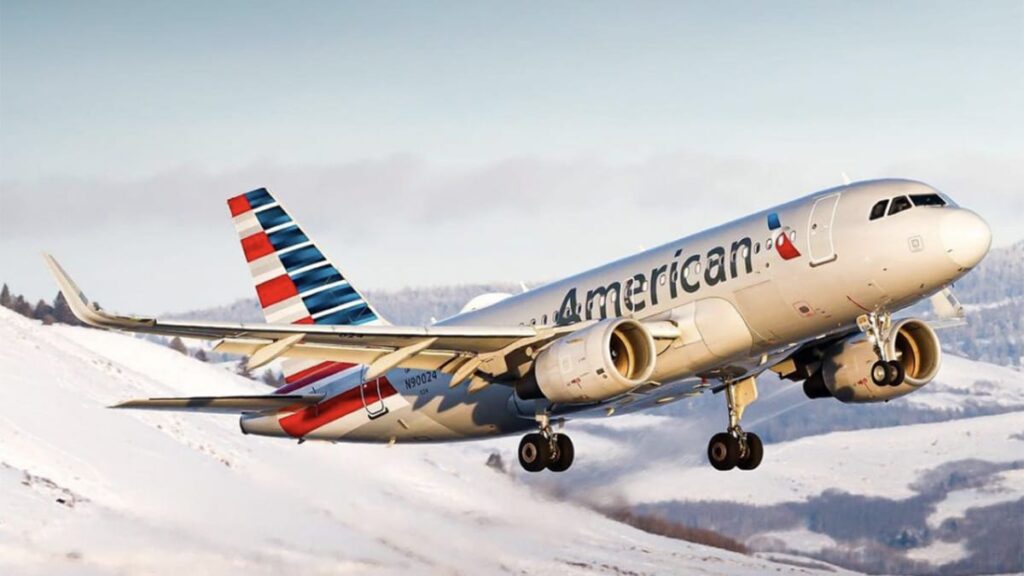 American Airlines jet taking off over a snow-covered landscape with a blue sky and distant hills in the background