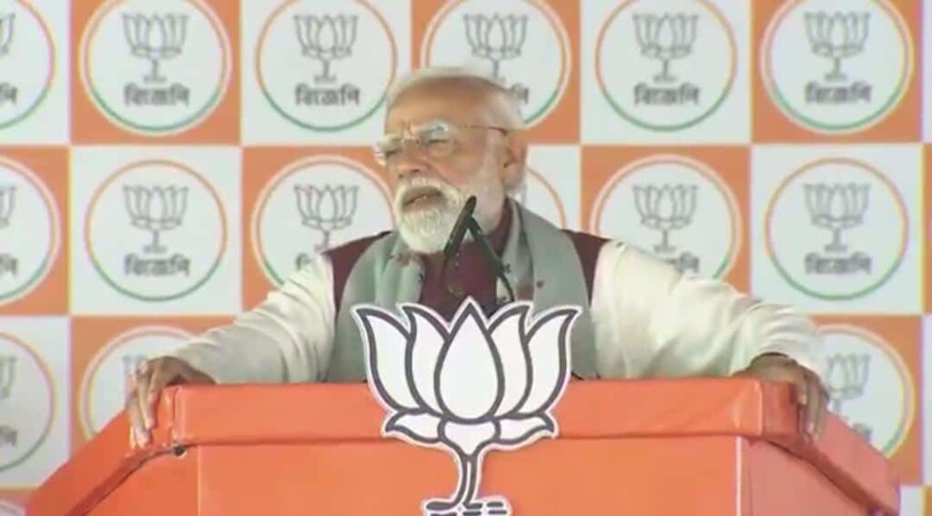 Bearded man with glasses speaking at a podium with an orange-and-white BJP lotus emblem backdrop and logo on the front