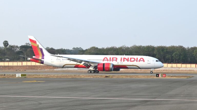 Air India wide‑body airplane on the taxiway with red logo, white fuselage, and a colorful tail at the airport ground.