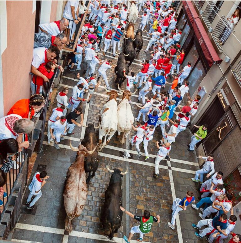 Crowd of runners in white with red scarves in a narrow street as bulls pass through, surrounded by excited spectators on balconies and sidewalks.