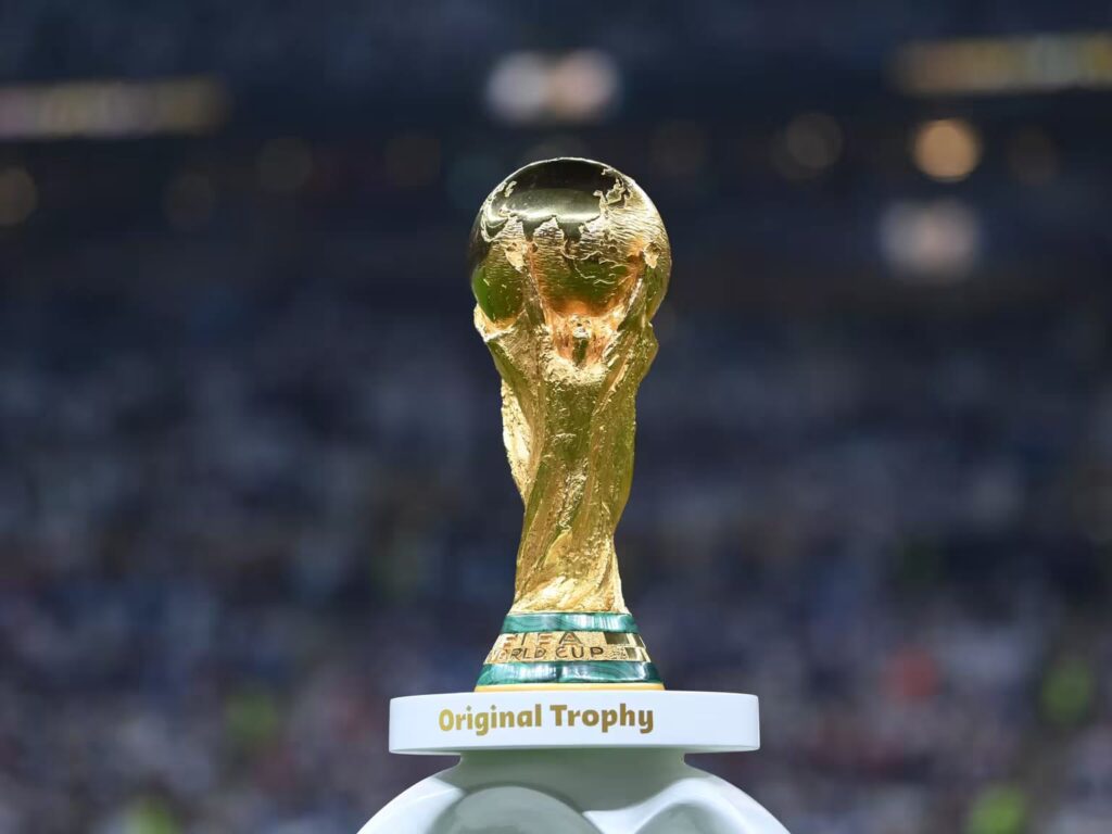 Gold FIFA World Cup trophy on a pedestal with 'Original Trophy' label in a stadium backdrop