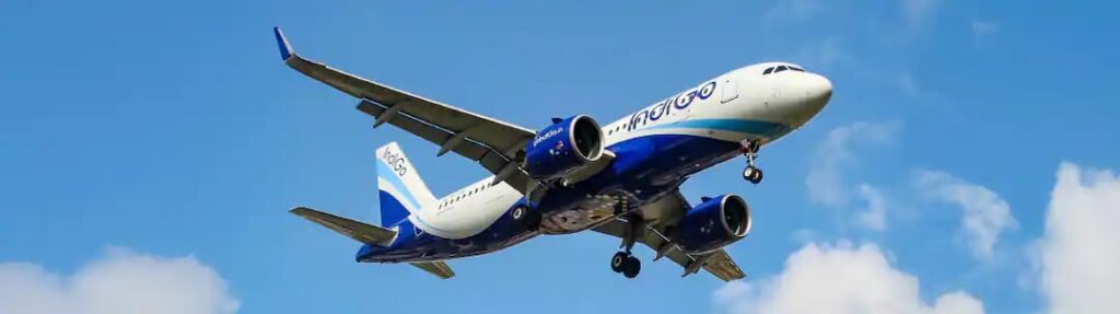 Blue-and-white commercial airliner in flight with landing gear down against a clear blue sky with clouds nearby.