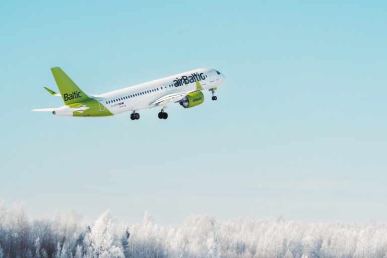 Air Baltic passenger jet taking off over snowy treetops with a pale blue sky background.