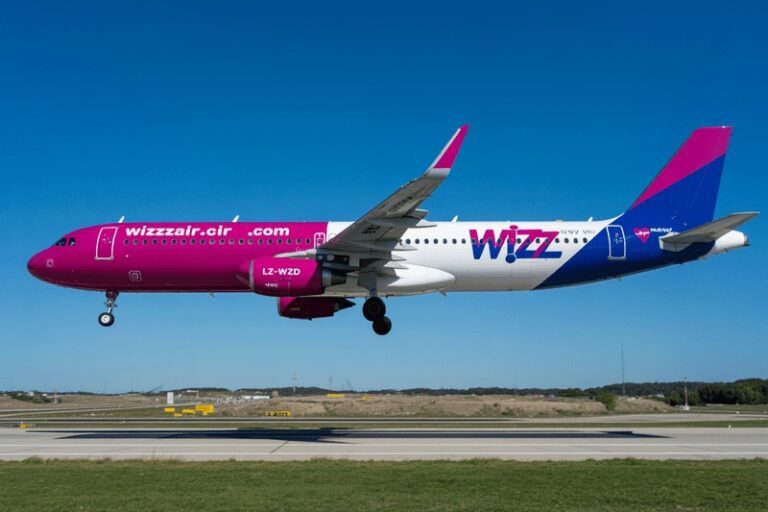 Wizz Air passenger jet in magenta, white, and blue livery in flight above a runway.