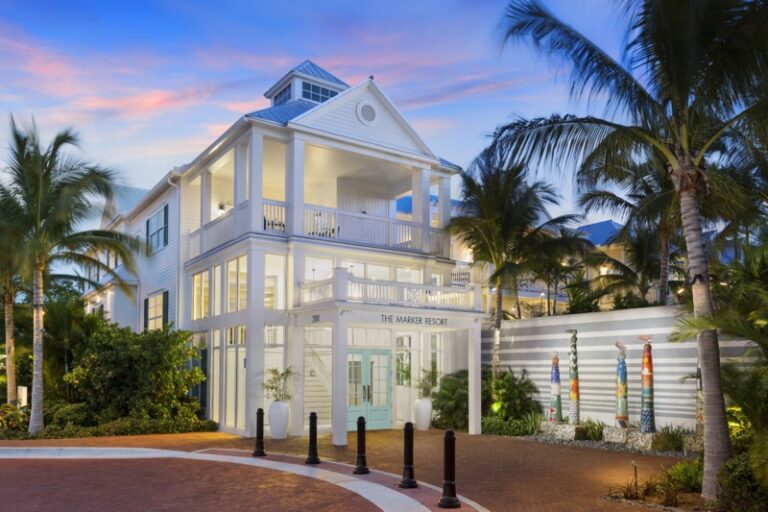 Three-story white coastal resort called The Marker Resort, with balconies, blue doors, and palm trees at sunset.