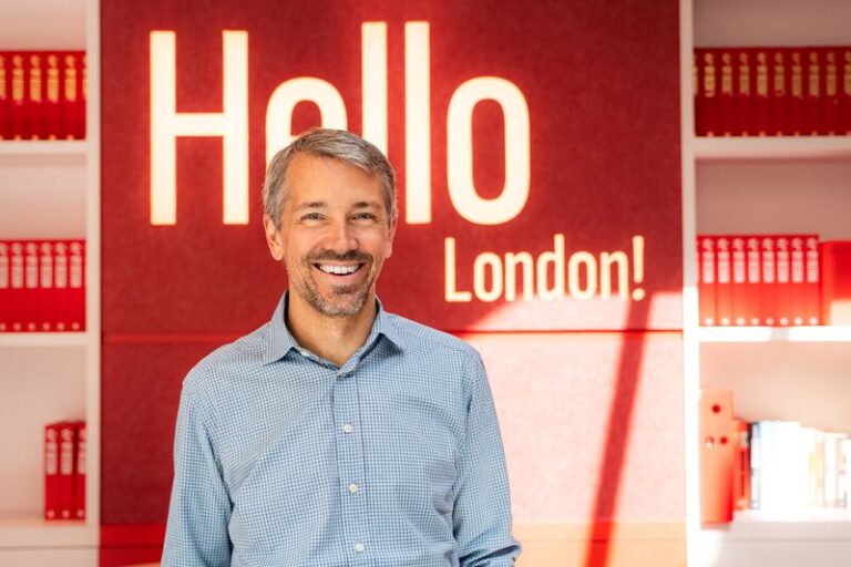 Smiling man in a blue checkered shirt stands in front of a red wall with 'Hello London!' sign behind him.