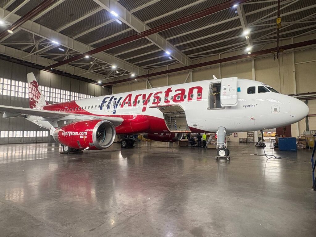 FlyArystan jet in a maintenance hangar with its passenger door open and bright overhead lights