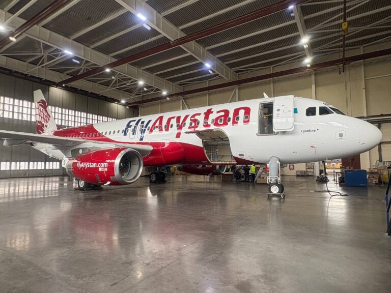 FlyArystan jet in a maintenance hangar with its passenger door open and bright overhead lights