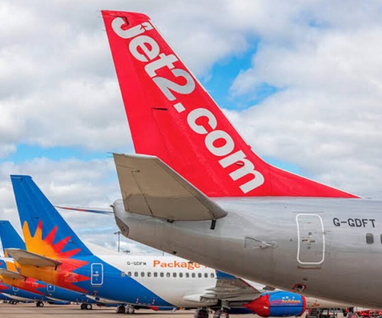 Rear view of a Jet2.com airplane with a bright red tail bearing the Jet2.com logo, parked on an airport tarmac with other aircraft in the background.