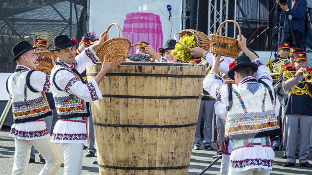 People in traditional embroidered outfits lift baskets of grapes toward a large wooden barrel at a harvest festival, with a brass band nearby.