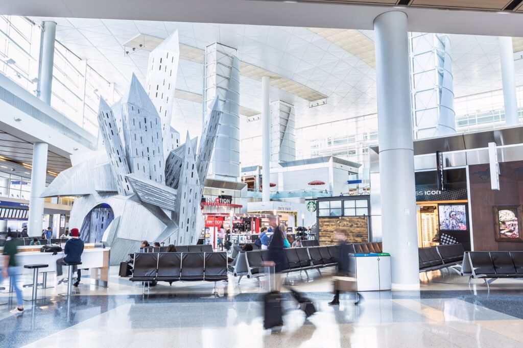 Interior of a bright, modern airport with a large geometric metal sculpture, seating area, and various shops in the background.