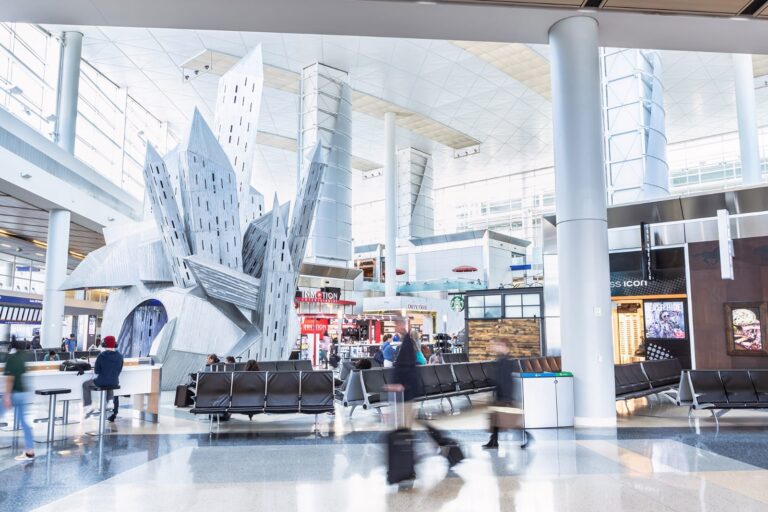 Interior of a bright, modern airport with a large geometric metal sculpture, seating area, and various shops in the background.