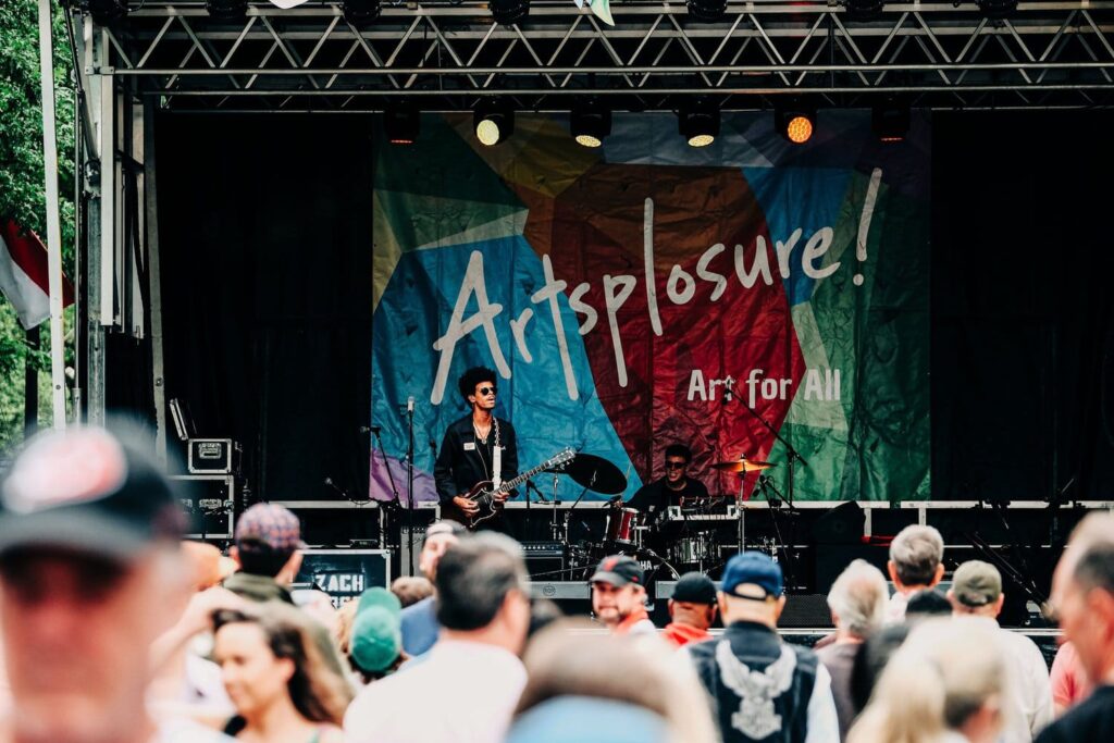 Live band performing on an outdoor stage with a colorful banner reading 'Artsplore! Art for All' and a cheering crowd in the foreground.