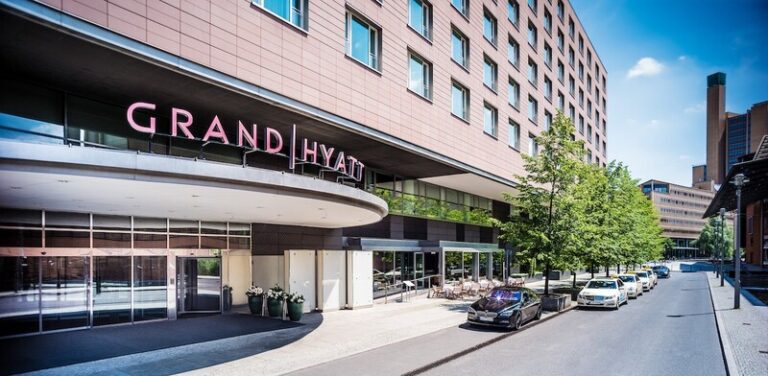 Grand Hyatt hotel entrance on a sunny city street with glass doors, a pink sign, outdoor seating, trees, and parked cars.