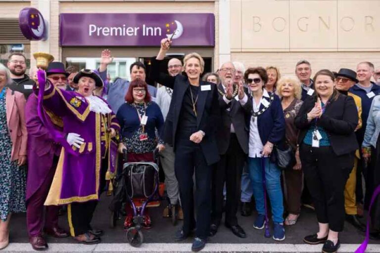 Group of people posing for a photo at a Premier Inn opening, with several smiling attendees and one person in a purple ceremonial outfit waving in front of the sign.