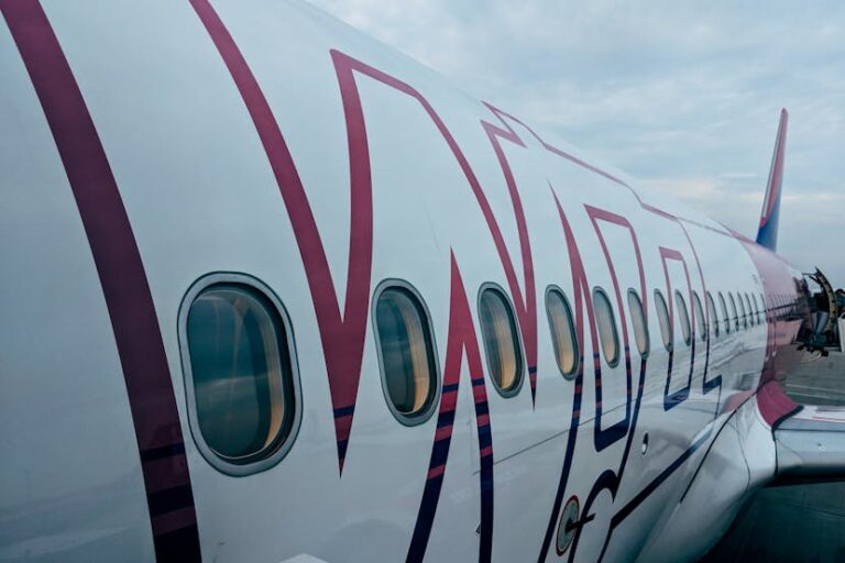 Side view of a passenger airplane with magenta branding on the tarmac, showing rows of windows and the fuselage. Readable branding along the side is visible into the distance.