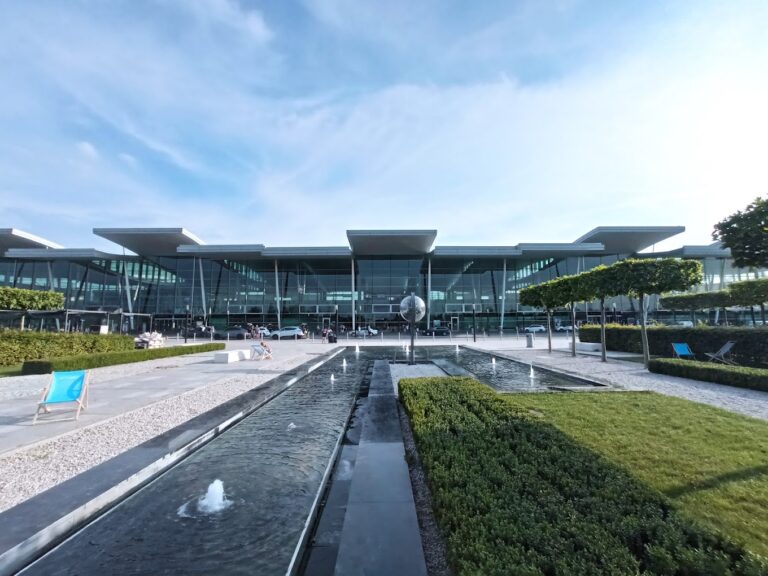 Modern glass-front building with a central entrance, reflecting pool, and neatly trimmed hedges in a sunny plaza.
