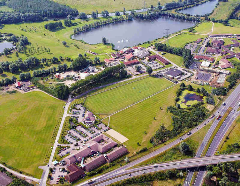 Aerial view of a campus-like complex with red-roof buildings, expansive green fields, a large lake, and a nearby highway.