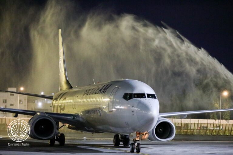 Nighttime view of a large commercial jet on the taxiway with a misty exhaust plume behind it and a Philippine Department of Tourism seal in the foreground.