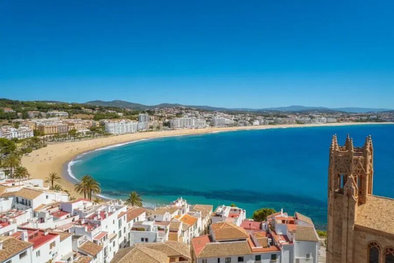 Aerial view of a sunny coastal town with a curved sandy beach, turquoise bay, white buildings, and a church tower on the right.