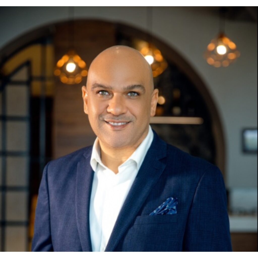 Portrait of a smiling bald man in a navy blazer and white shirt, standing in a modern office setting with warm lighting behind him.