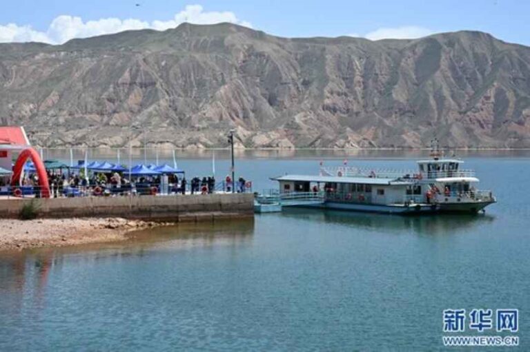 People gathered at a lakeside dock with blue tents, a passenger boat on the water, and rocky mountains in the background