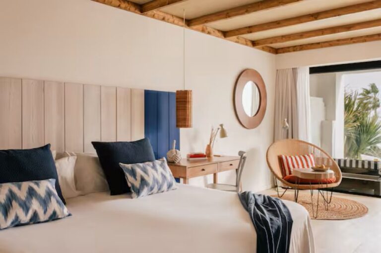 Bright hotel bedroom with a large bed, blue accent pillows, and a wooden desk by a round mirror and wicker chair by a glass door to the outdoors.
