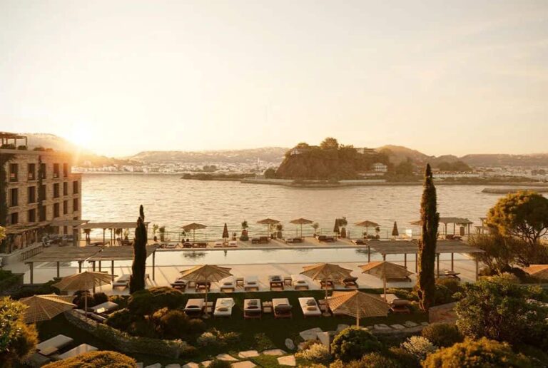 Sunset over a resort pool area with loungers, umbrellas, and a calm sea in the background, island visible on the horizon.