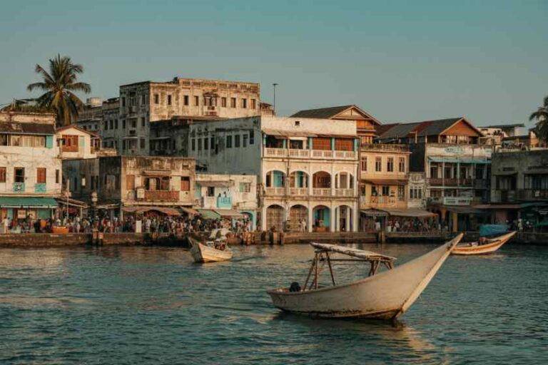 Boats sail on a calm harbor with pastel, colonial-style buildings lining the waterfront market. In the foreground, a wooden boat glides past a lively quay.
