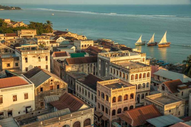 Coastal town with old yellow and beige buildings by the water, and three sailboats on the bay.