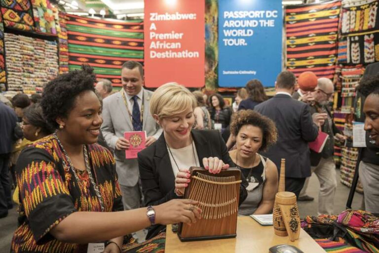 Women at a cultural expo examining a wooden mbira (thumb piano) with colorful fabrics around and Zimbabwe banners in the background.