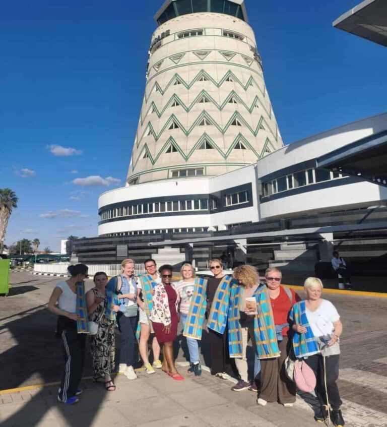 Tour group posing outside a tall, cylindrical tower with green zigzag windows; everyone wearing blue-and-yellow scarves.