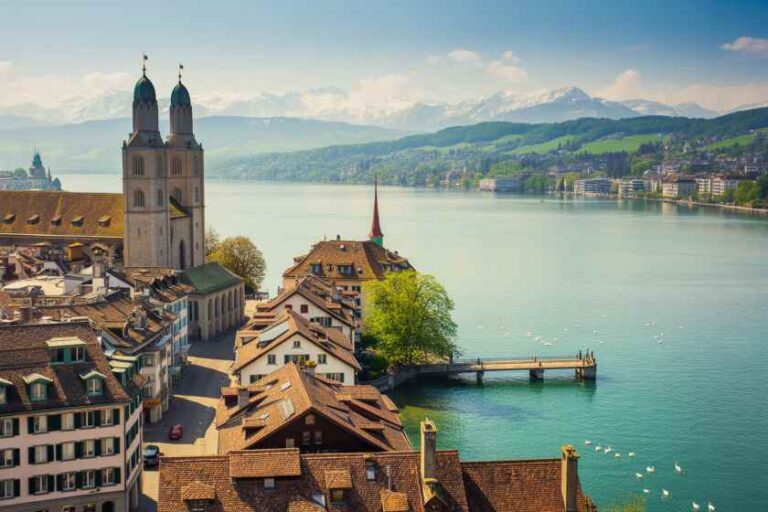 Aerial view of a European lakeside town with red-tiled roofs, a two-towered church, and a calm lake with green hills and mountains in the distance.