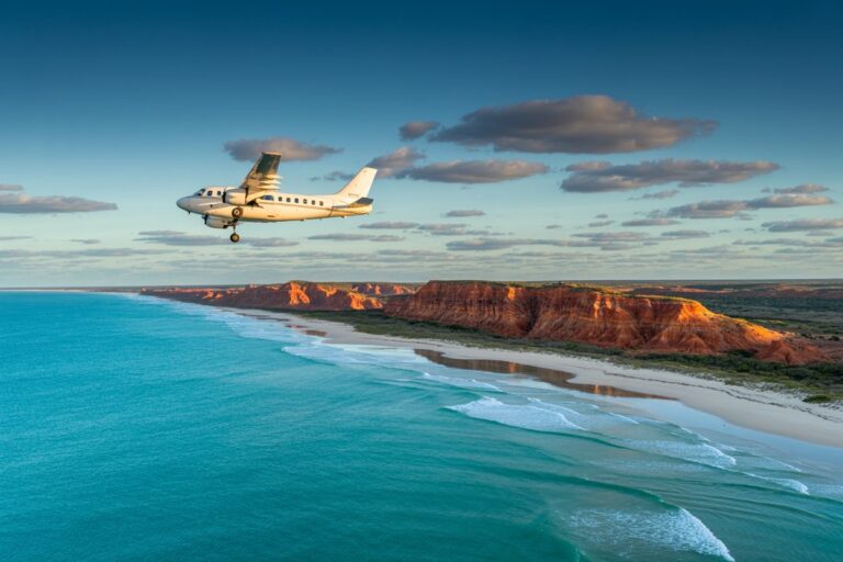 Small white airplane flying over a turquoise ocean toward a red rocky coastline and white-sand beach.