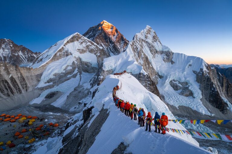 Line of climbers in bright jackets trekking along a snowy ridge with prayer flags fluttering, cliffs and tents visible below.