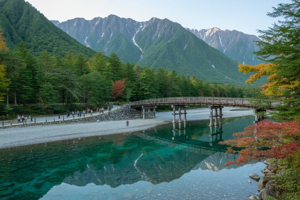 Wooden bridge spans a clear turquoise river; people walk along the pebble shore with a forest and rugged green mountains in the background.