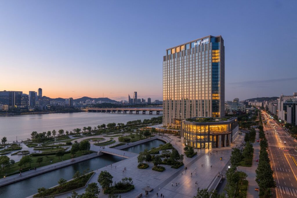 Evening cityscape by a river with a tall glass building lit gold on the right and a landscaped waterfront park in the foreground.