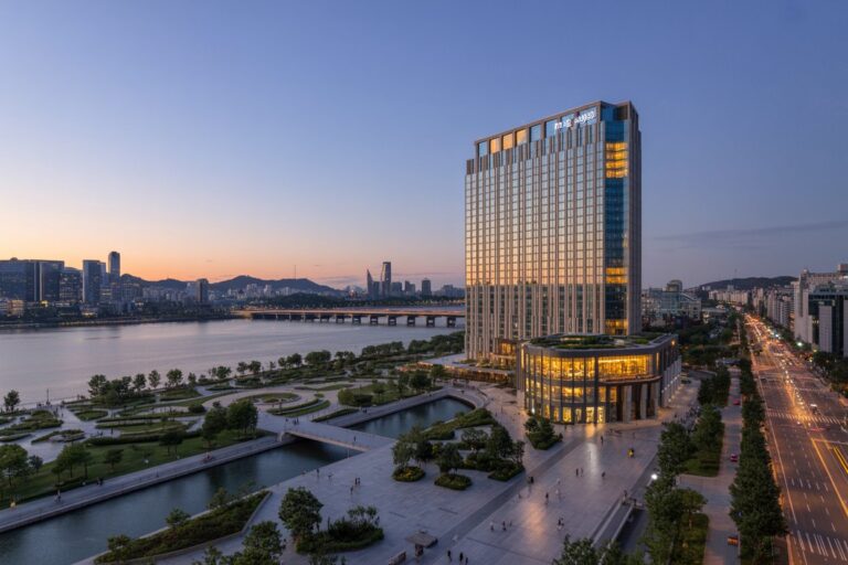 Evening cityscape by a river with a tall glass building lit gold on the right and a landscaped waterfront park in the foreground.