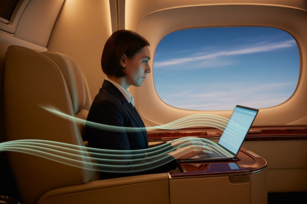 Businesswoman in a suit typing on a laptop while seated by an airplane window, blue sky outside.