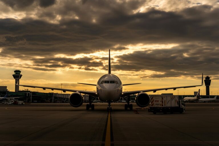 Front view of a large commercial jet on the runway at sunset, airplane lights off, with ground service vehicles nearby and control towers in the background