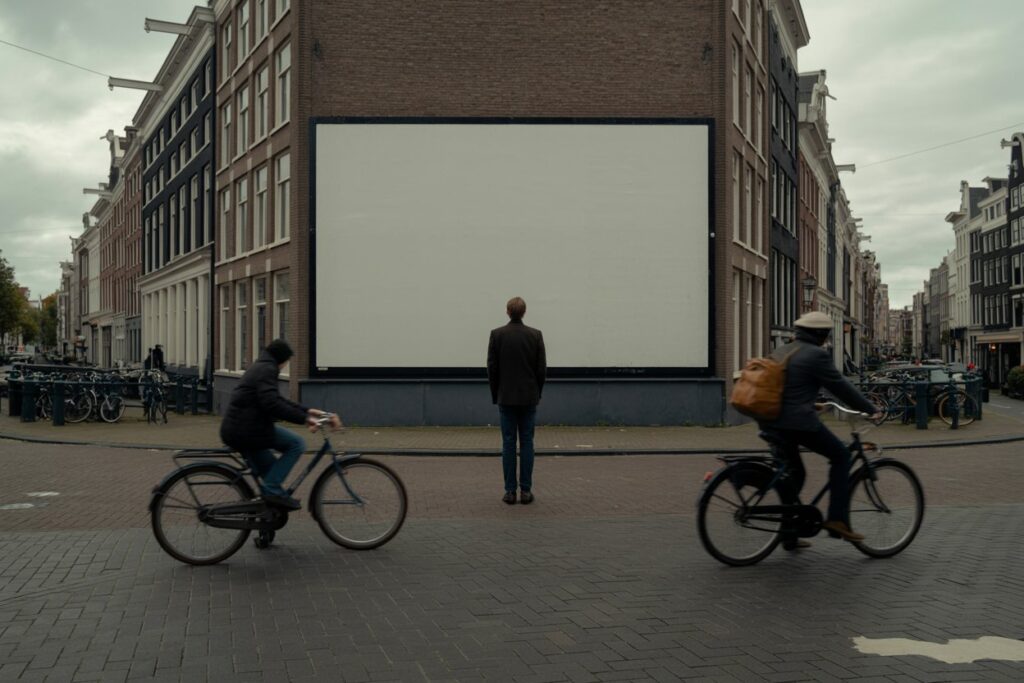 Man stands alone facing a large blank billboard on a brick building in a city street, while two cyclists ride by in the foreground.