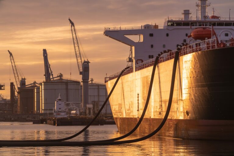Large cargo ship moored at a dock at sunset, with thick mooring lines curving along the hull.